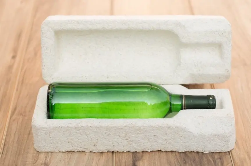 A green glass bottle is securely positioned within a custom white foam insert inside an open packaging box, resting on a wooden surface.